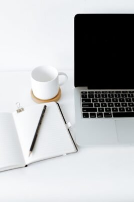 A laptop on a white table, with a mug and a pencil resting on an open notebook on the left side.
