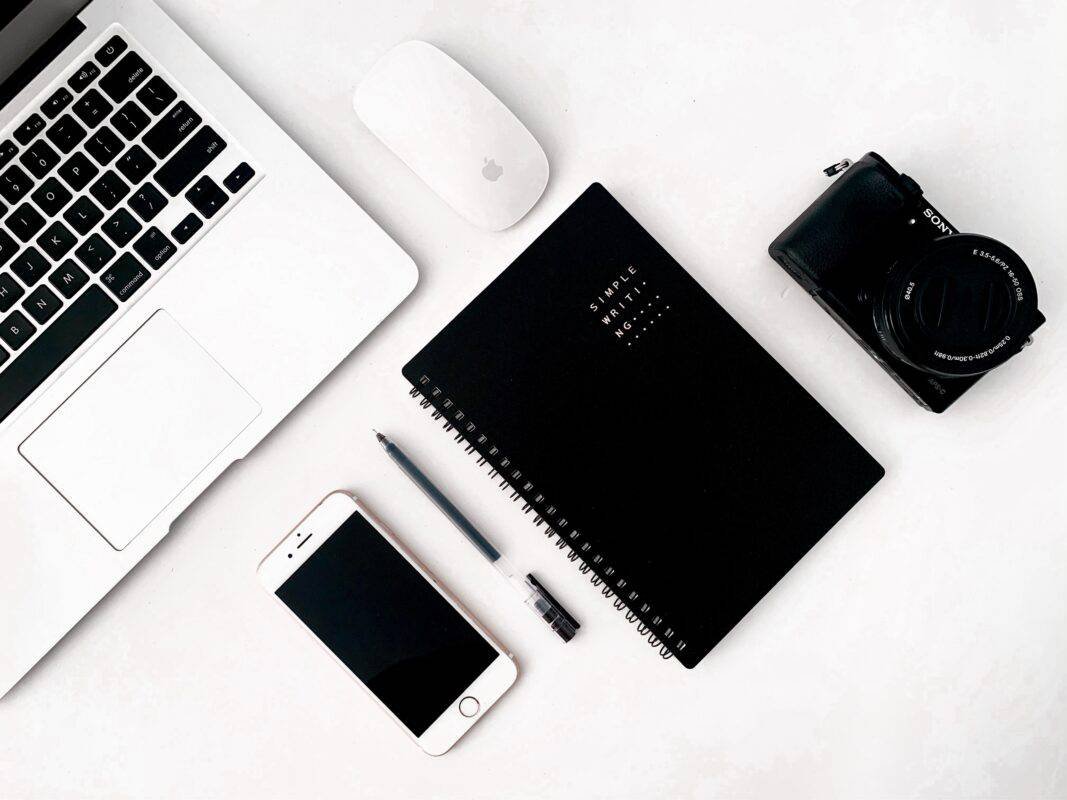 A portion of an Apple laptop and mouse can be seen sitting on a table. In front of them are a cell phone, pen, notebook, and a Sony camera.