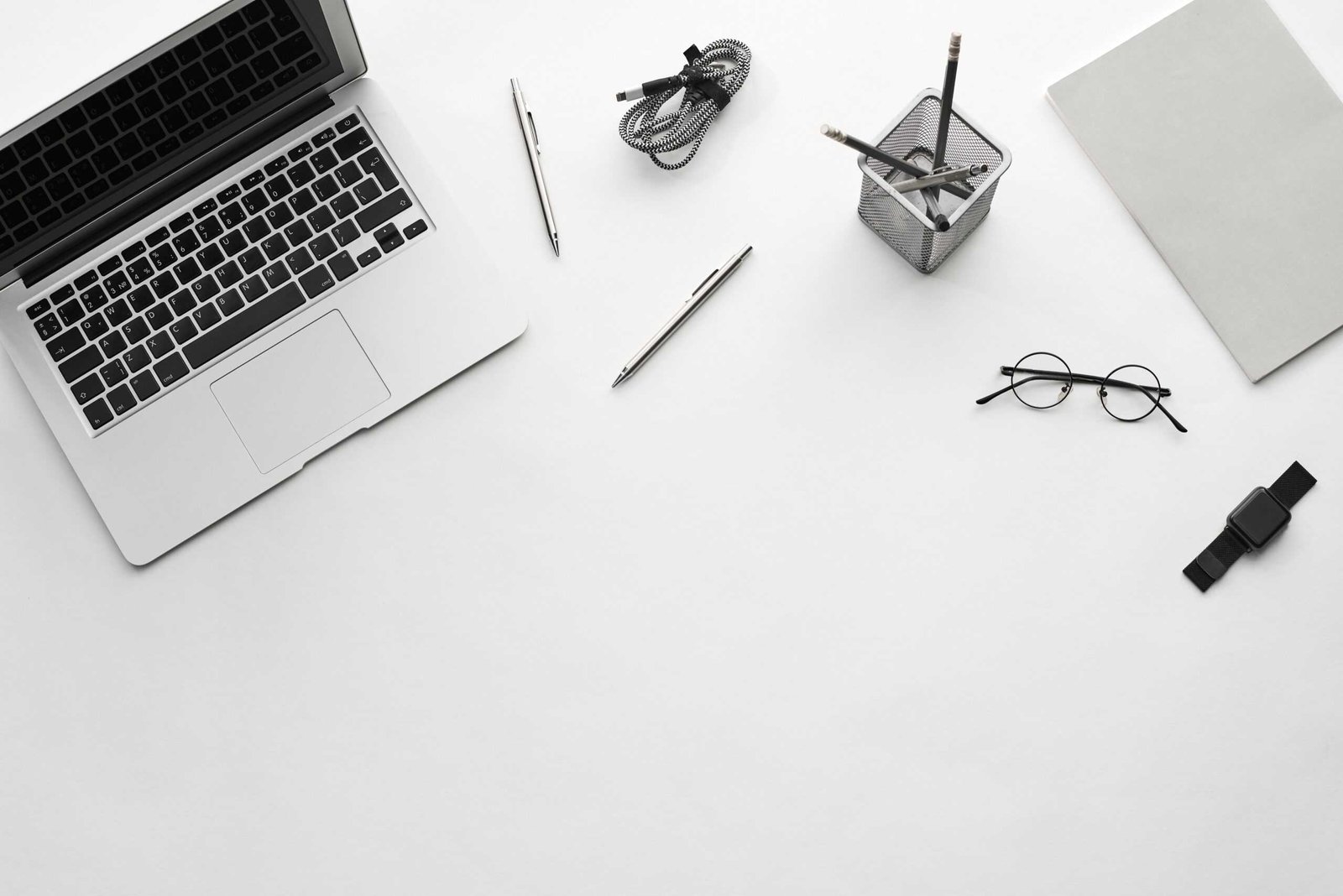 A laptop sitting on a white table, with pens, pencils, a connector, eyeglasses, a black watch, and a notebook nearby.