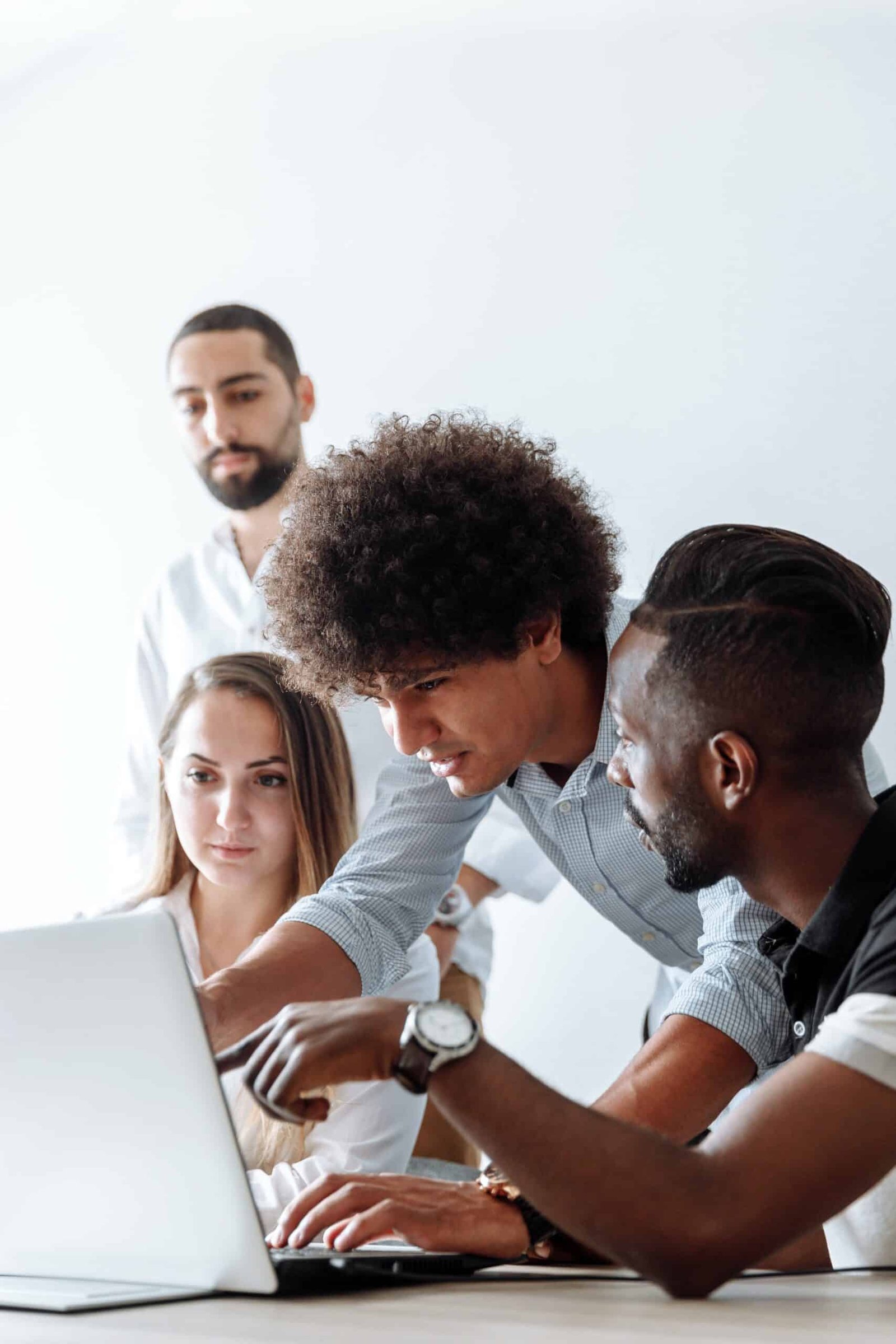 Four individuals engaged in collaborative discussion, with one standing at the back, one bent toward the laptop, and the other two sitting beside the table. All are looking intently at the laptop screen as they converse.