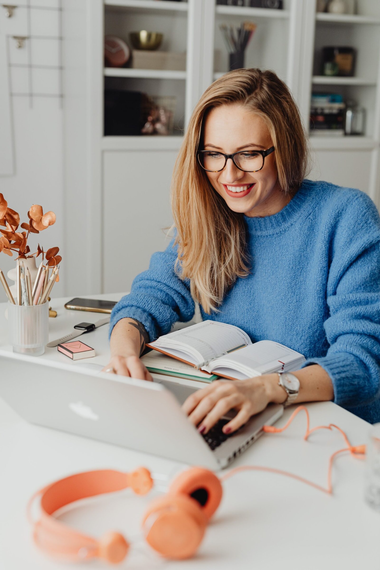 A woman in a blue sweater, wearing eyeglasses and a watch, is focused on her laptop, with pens and notebooks in front of her.