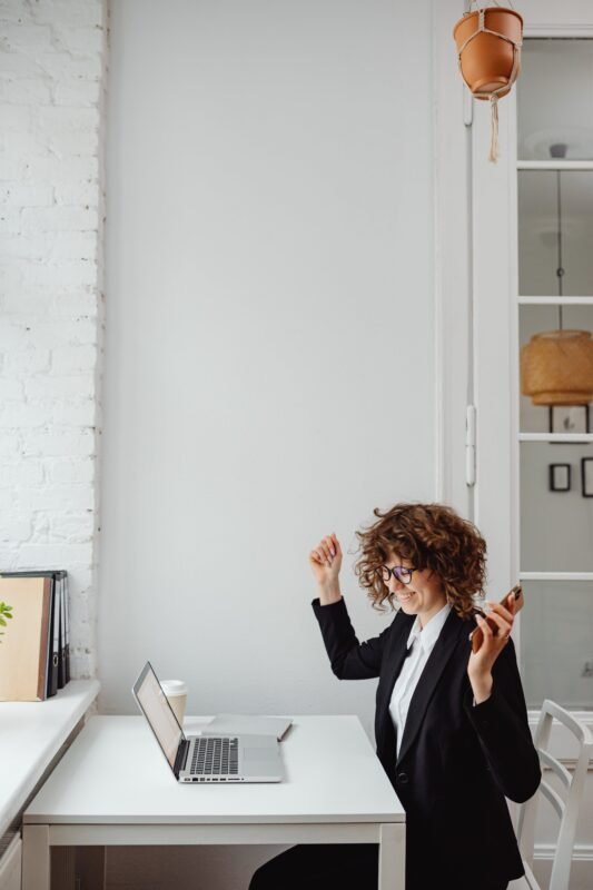 A curly-haired woman in a black suit, wearing glasses, happily raises both arms in front of her laptop, left hand holding a cellphone. Nearby, a cup of drinks and a notebook are placed.