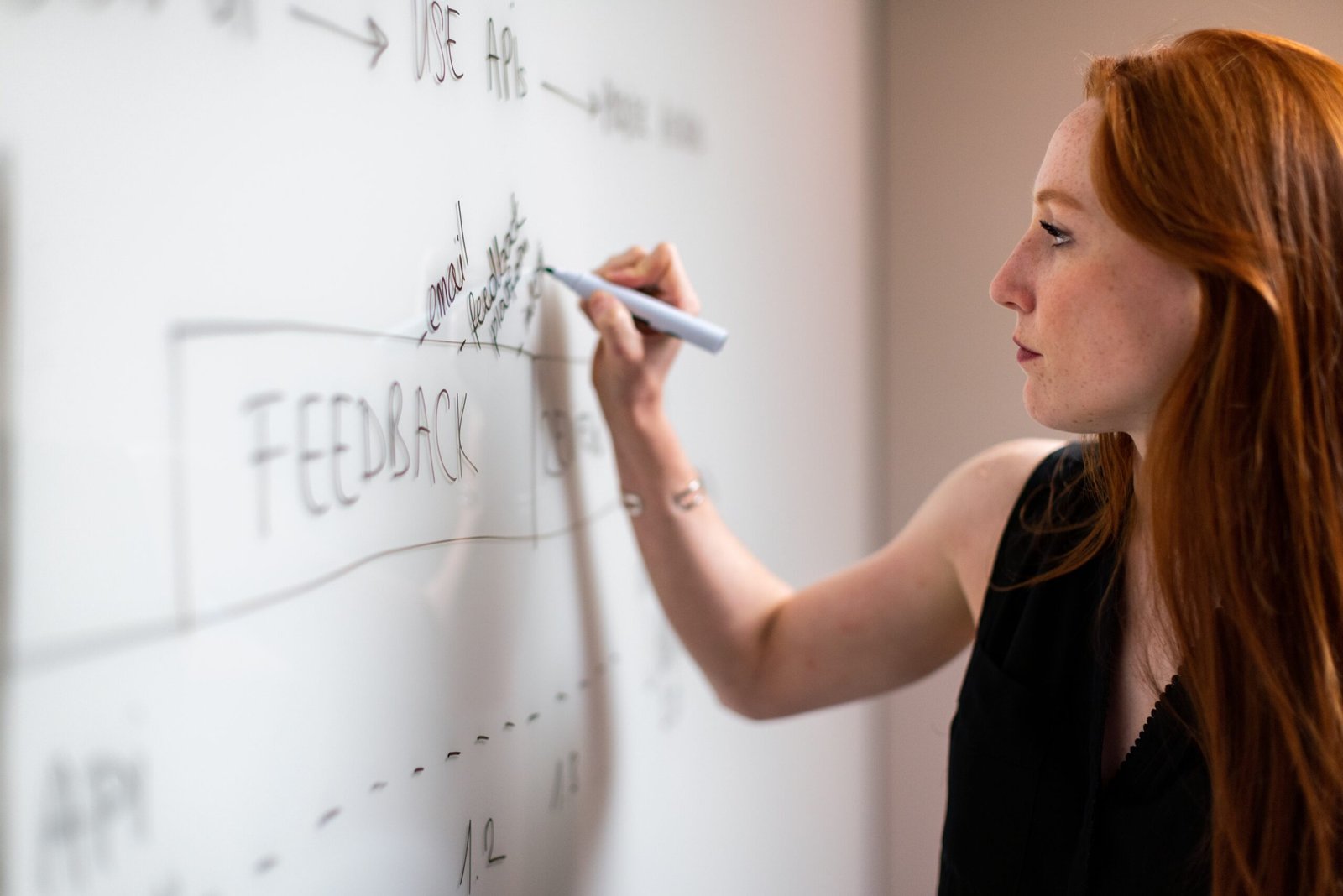 A long-haired woman in a black blouse is writing on a whiteboard.