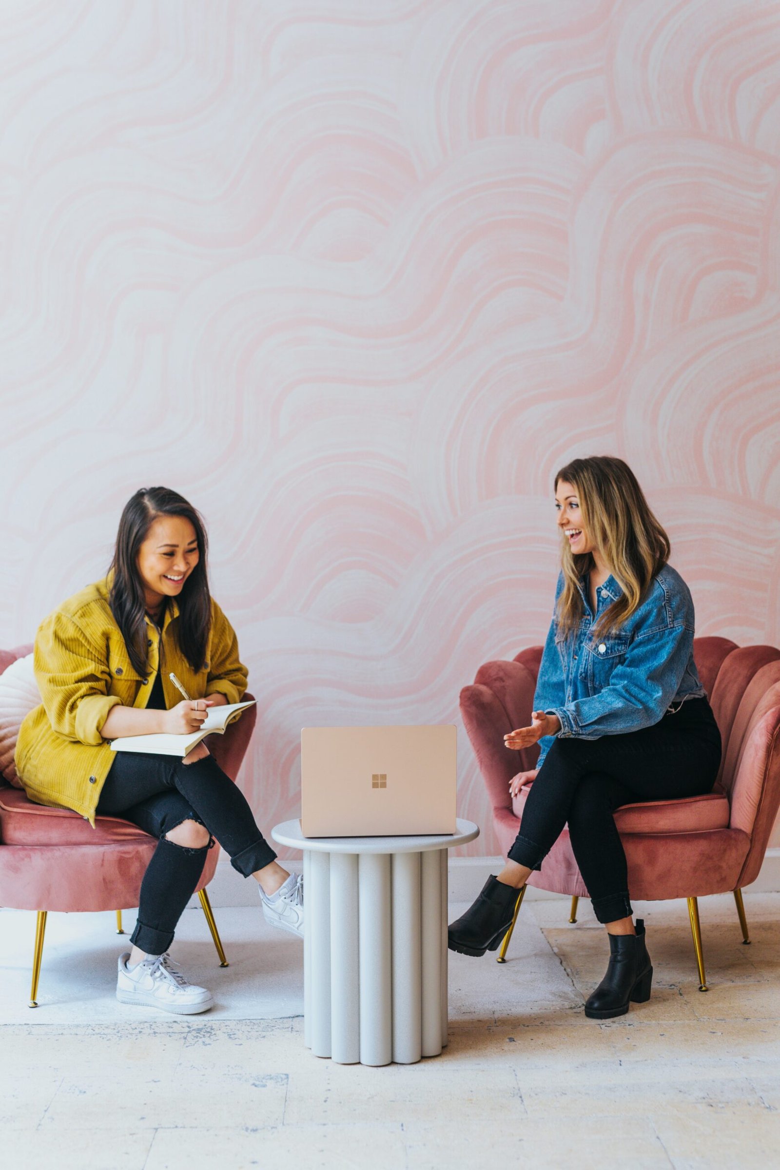 Two women engaged in discussion, one explaining while the other took notes. A laptop faced them as they conversed, with a pinkish mosaic-like design at their back.