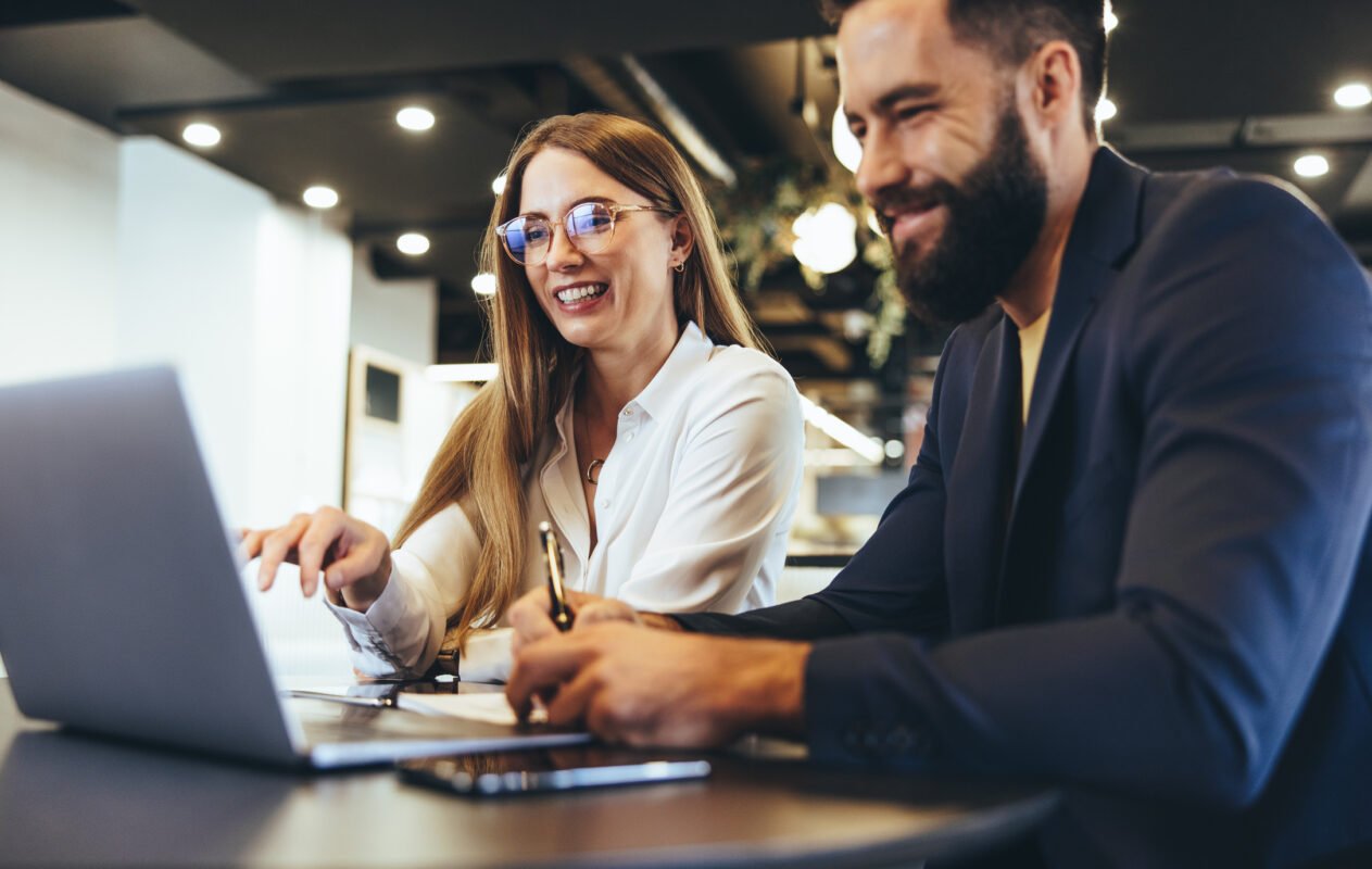 Two people engage in a conversation, with the man taking notes while the woman explains and scrolls on the laptop.