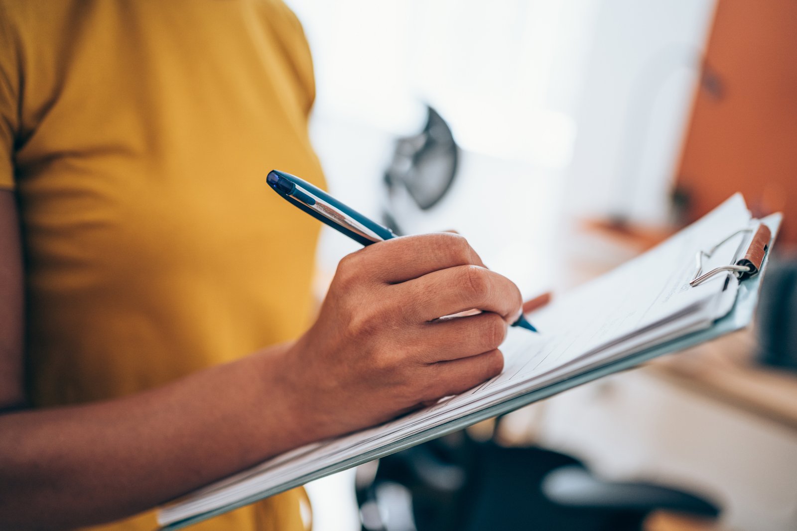 Cropped shot of a woman in orange clothing making notes on a clipboard inside an office.