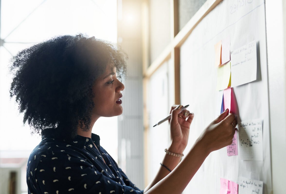 A curly-haired woman looks at a note pasted on a wooden wall while holding a pen in her raised left hand, with other pasted notes nearby.