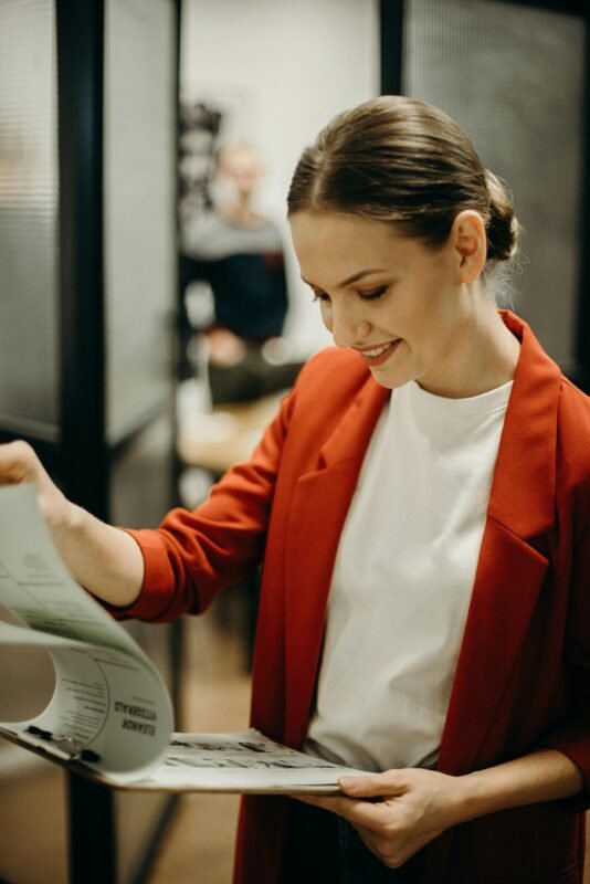 A woman in a red suit with a white blouse inside is enthusiastically reading something on a paper attached to a clipboard held in her left hand. Her other hand is holding the front pages.