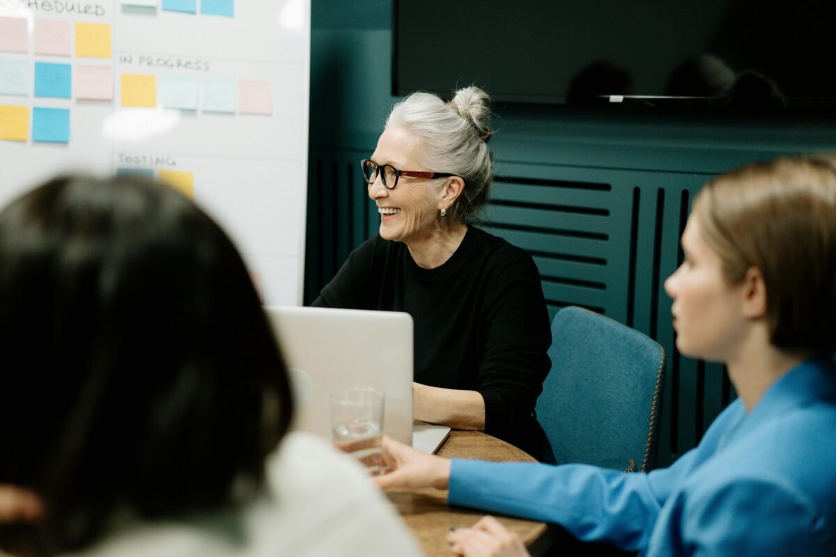 A group meeting focused on three individuals, sitting around the table. An old lady sat in front of the laptop, engaged in laughter while facing her colleagues, while another person held a glass of water. On one side of the room, a whiteboard adorned with sticky notes stood, adding visual aids to the discussion, while a flat-screen TV sat silent and off to the side.