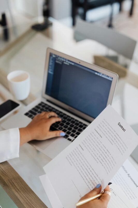 A woman is typing on her laptop while holding a paper contract in her other hand